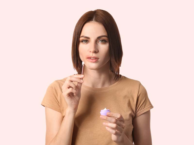 A woman with brown hear and a tan shirt is applying cream to a cold sore with a cotton swab