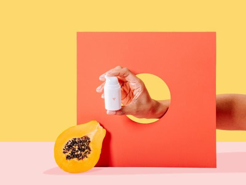 Woman's hand holding a bottle of Wisp Metronidazole Gel through an orange cardboard cutout with a cut open papaya on a yellow and pink background