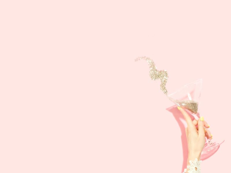 a woman's hand holding a martini glass pouring glitter onto a pink background