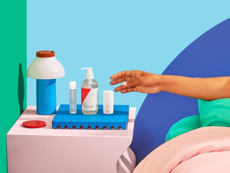 Woman's hand reaching for Wisp Balancing Wash, Harmonizing Lube, and OMG Cream on a pink nightstand with a blue lamp and green and blue walls