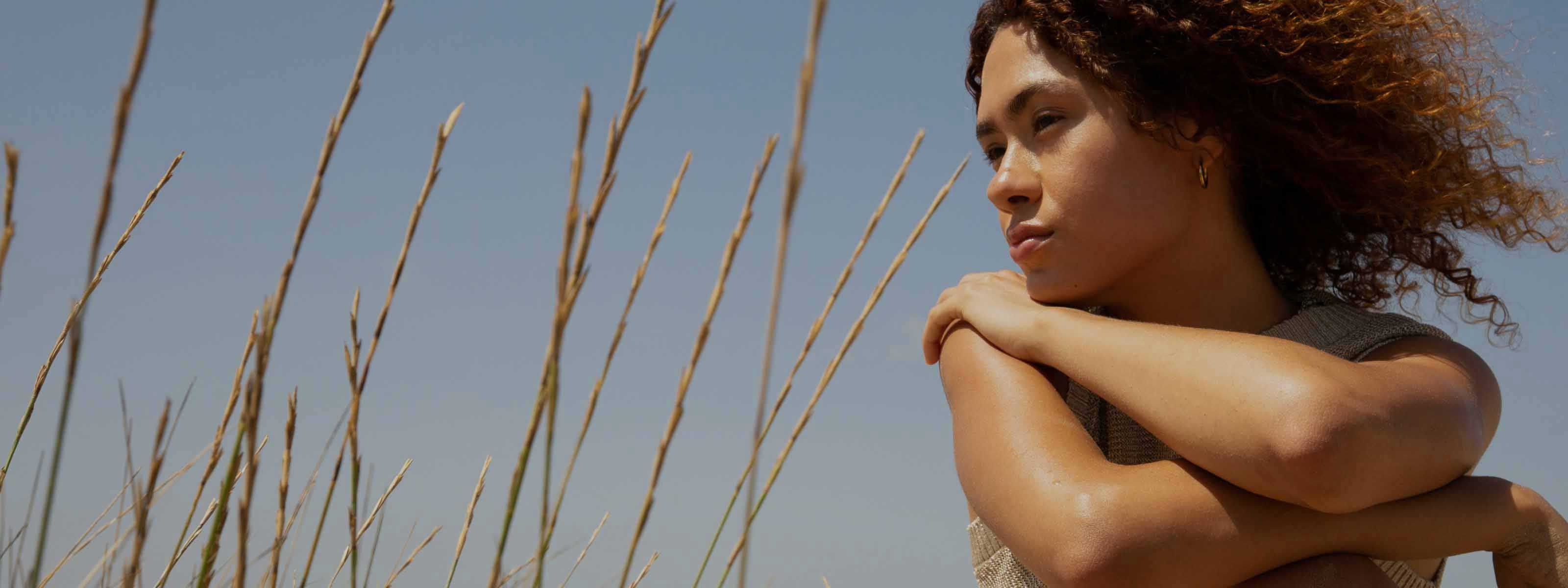 Person with curly hair resting arms on knees, looking into the distance in a field