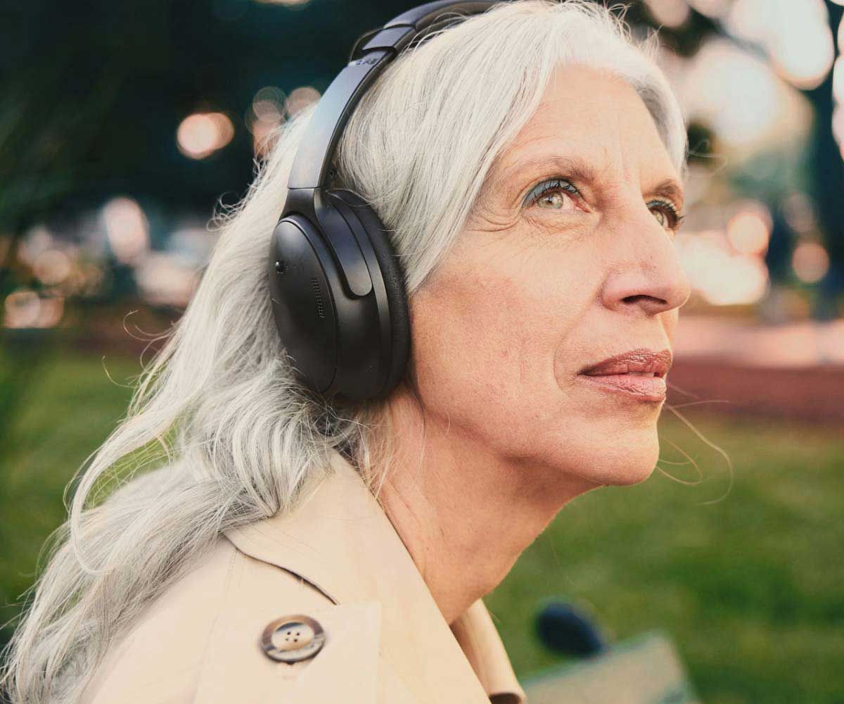 A close up of an older woman wearing headphones and a trench coat outdoors