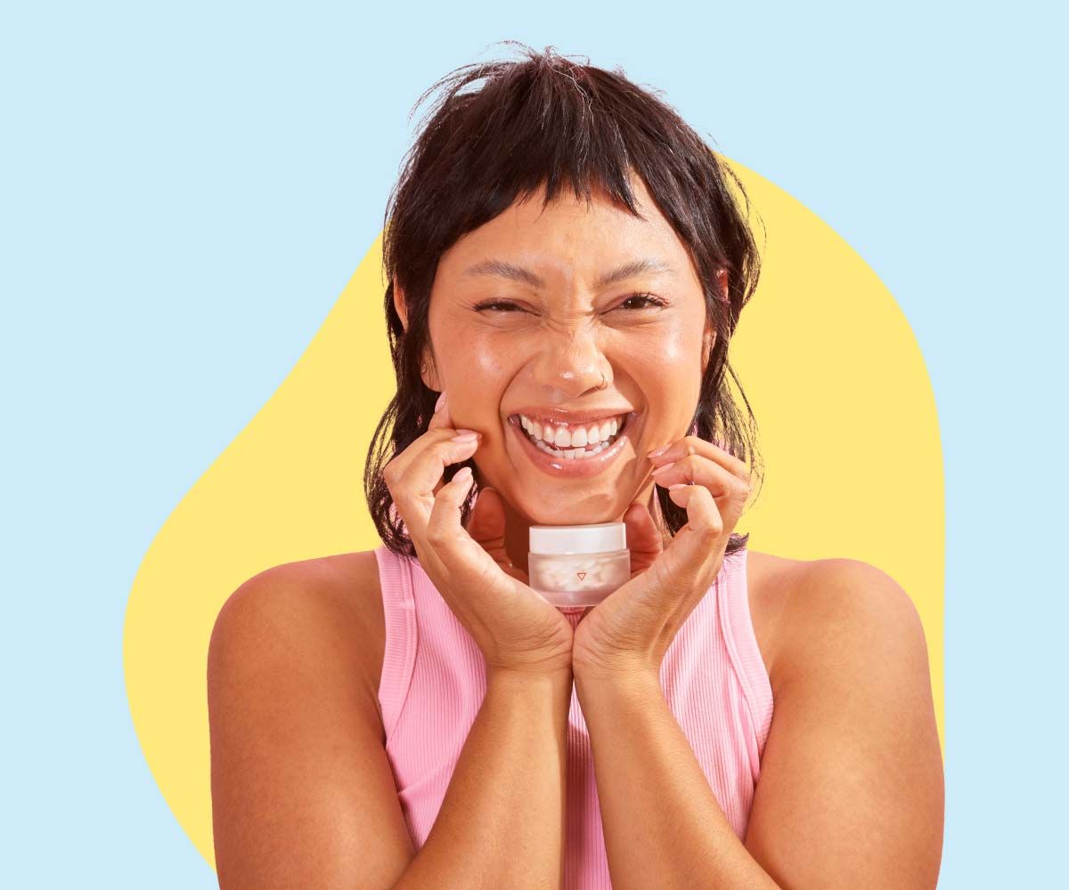 A smiling woman wearing a pink tank top is holding a Wisp glass jar under her chin in front of a light blue and yellow background