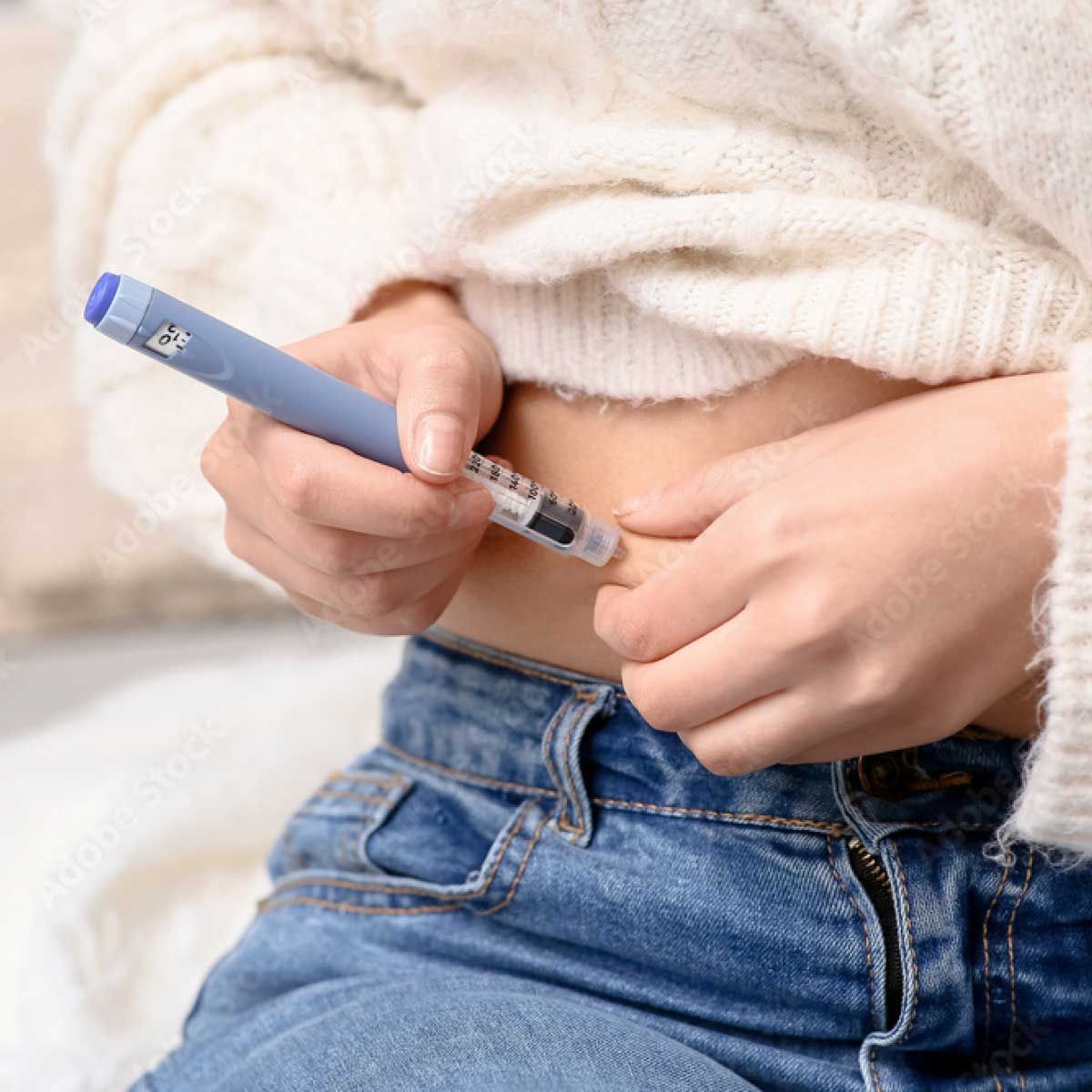 A person wearing a white sweater and jeans is self-administering an injection into their abdomen