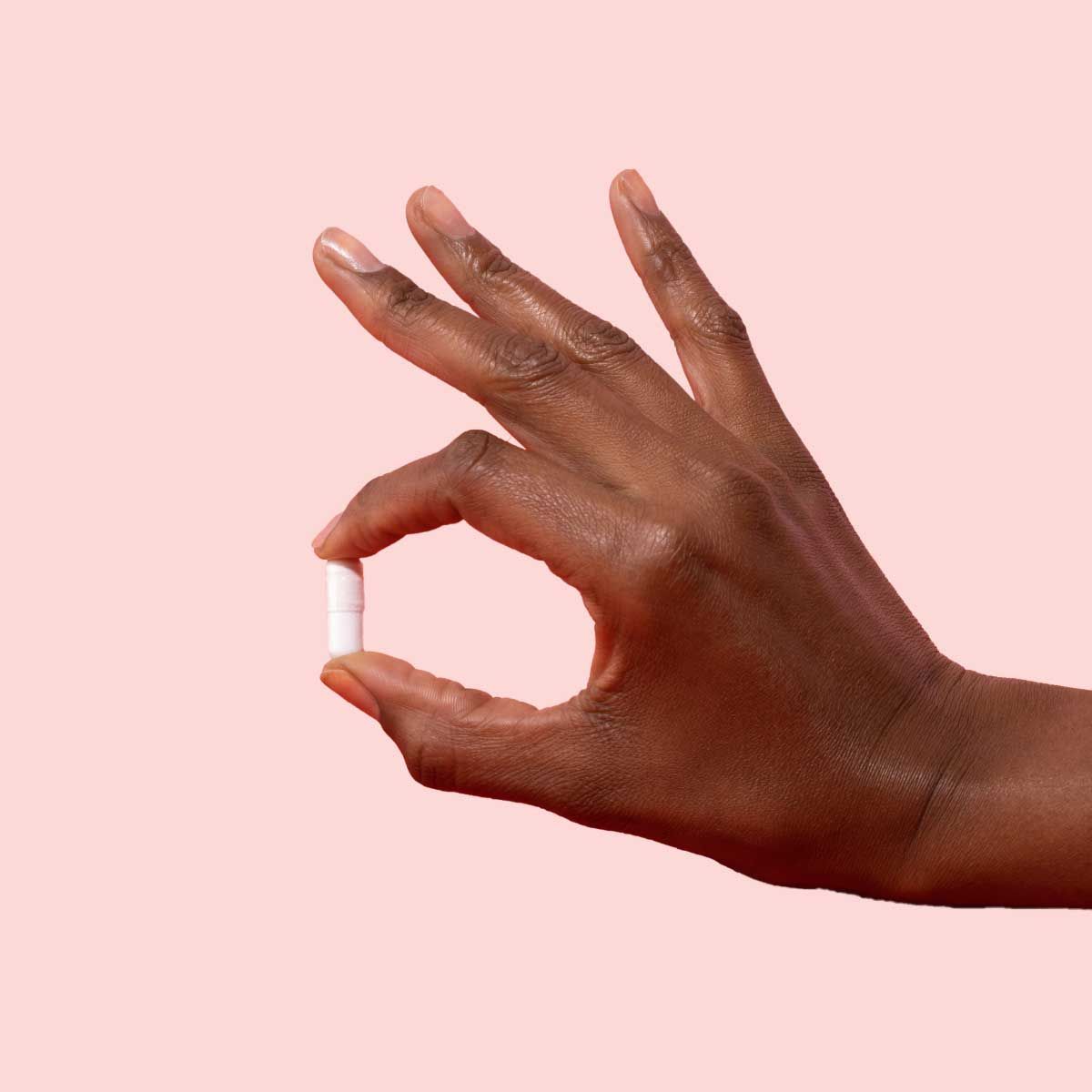 A person's hand holding a white capsule in front of a pink background