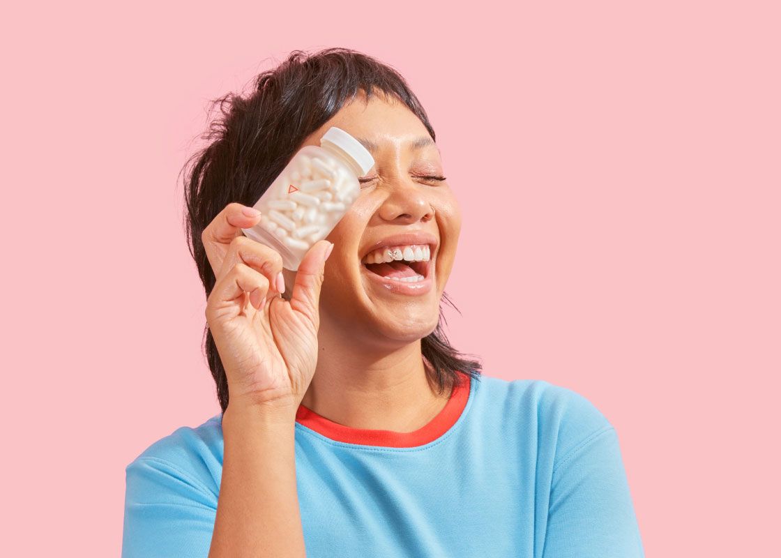 A woman smiling and wearing a red and light blue shirt while holding a Wisp glass pill jar in front of a pink background