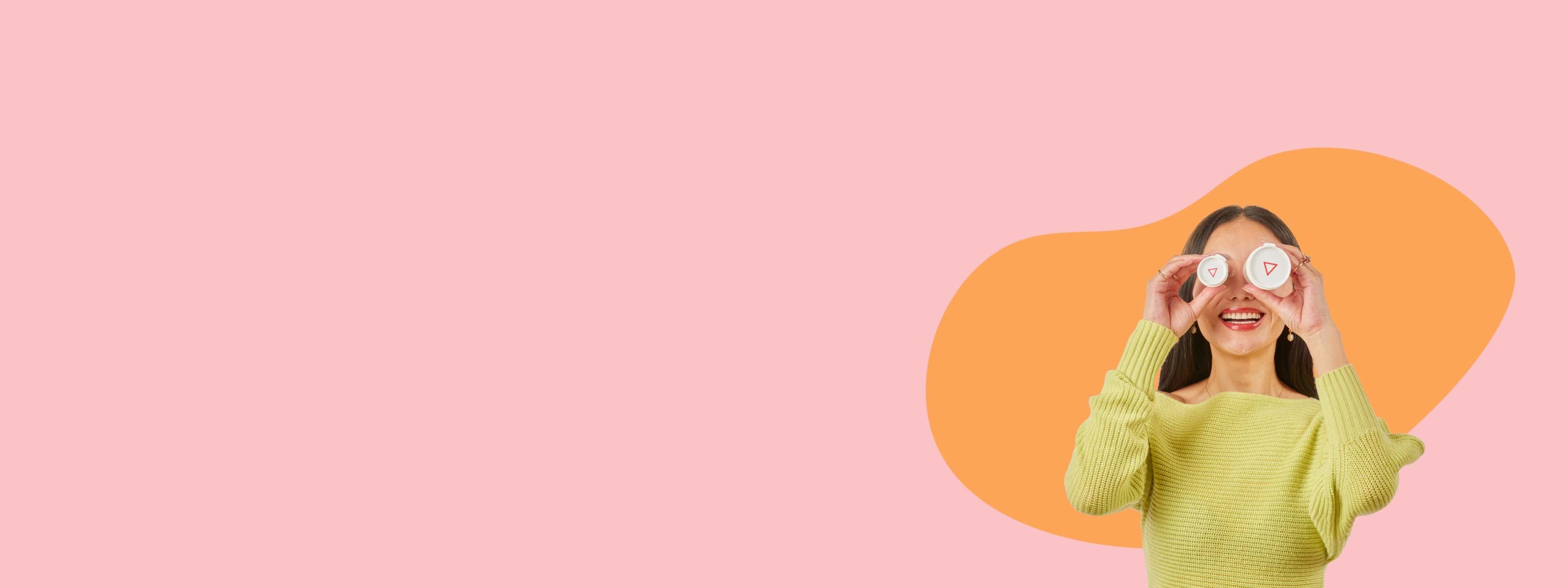 A woman smiling and wearing a bright yellow sweater is holding two Wisp bottles in front of her eyes in front of a pink background