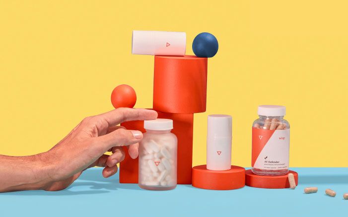 A man's hand reaching for a Wisp glass pill jar next to various other Wisp herpes treatment products balanced on colorful abstract shapes on a light blue surface with a yellow background