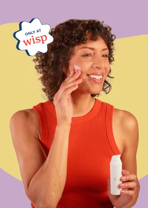 A smiling woman wearing a red tank top is apply Wisp Topical Spironolactone to her face