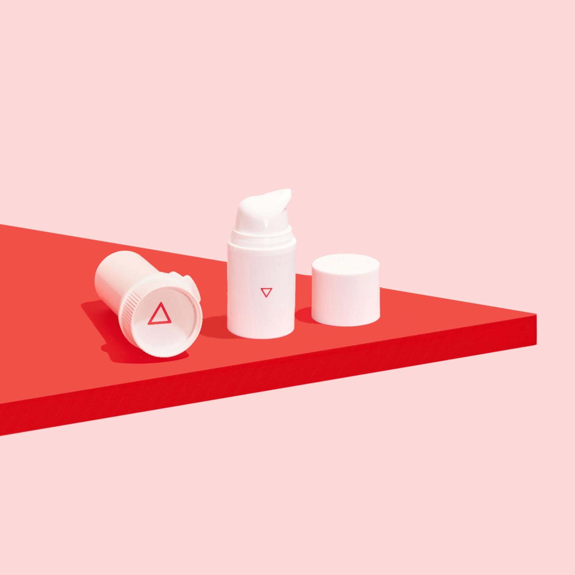 A pill vial laying on it's side next a pump bottle and lid on a red surface with a pink background