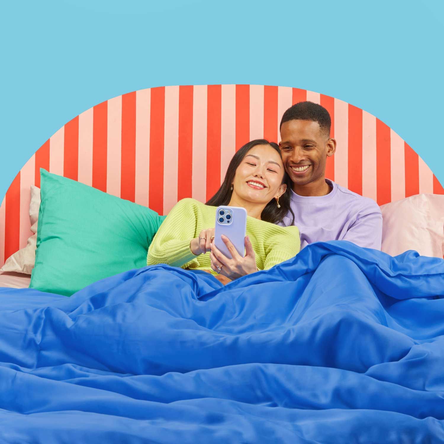 A couple laying in a colorful bed together smiling and looking at a mobile phone