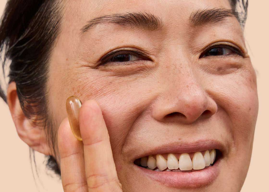 A close of a smiling woman holding a medication capsule with a cream background