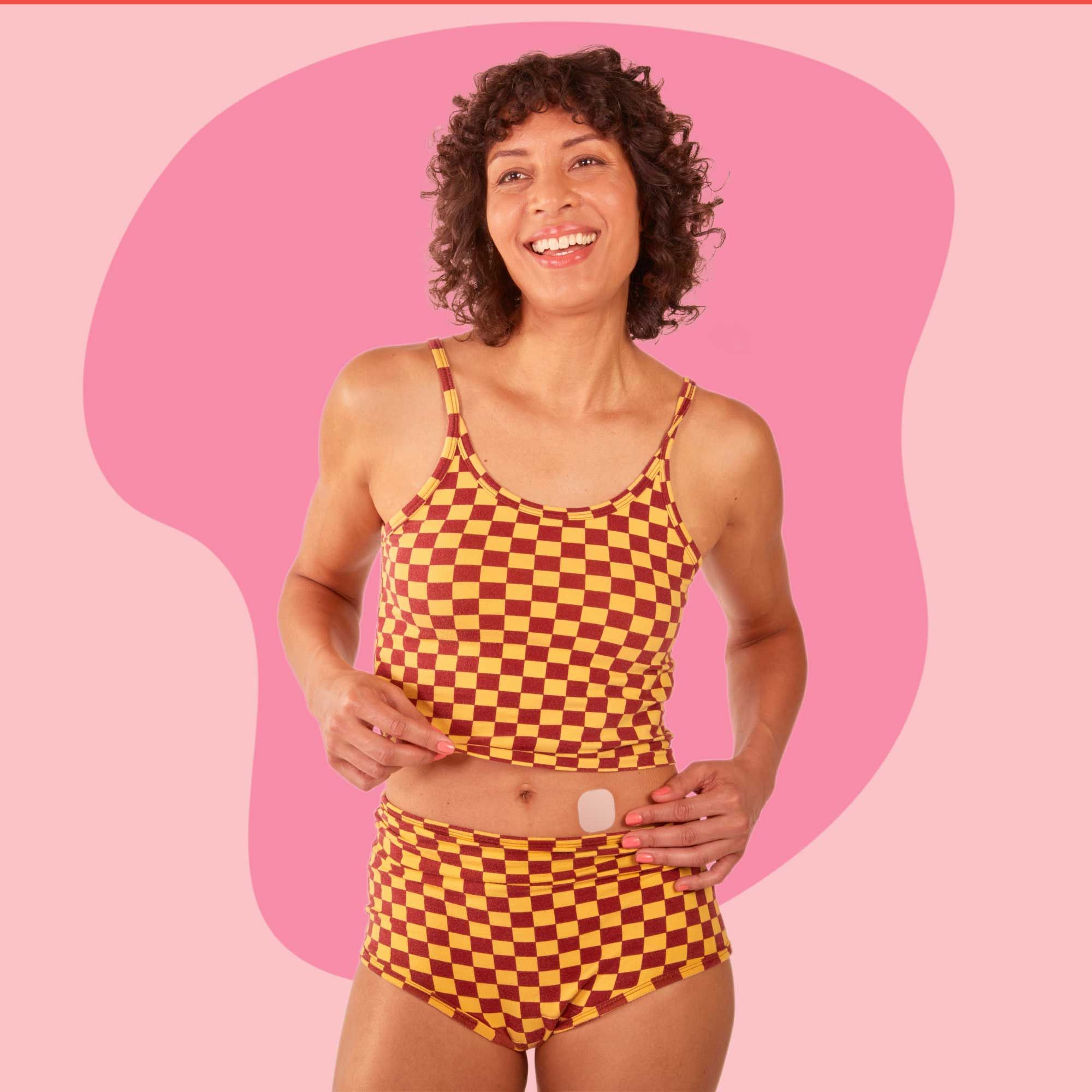 A woman wearing a checkered tank top and shorts is placing an estrogen patch on her stomach