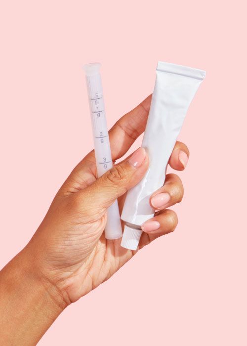 A woman's hand holding a tube of Estradiol Vaginal Cream and applicator in front of a pink background
