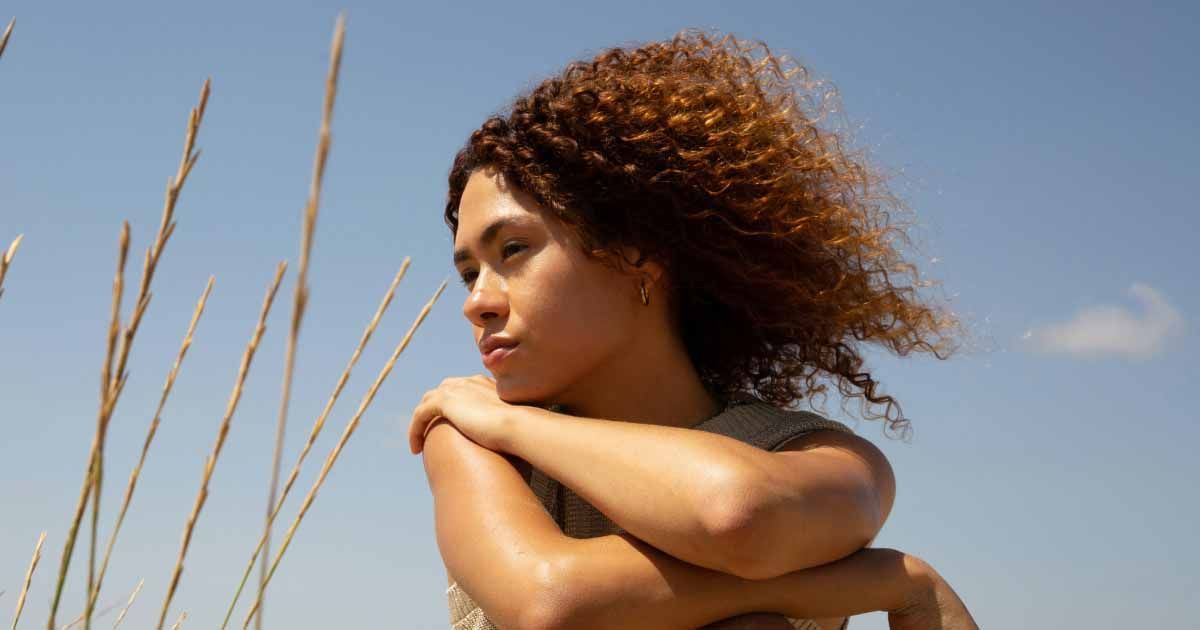 Woman with curly hair looking into the distance outdoors, with tall grass and a blue sky behind her.