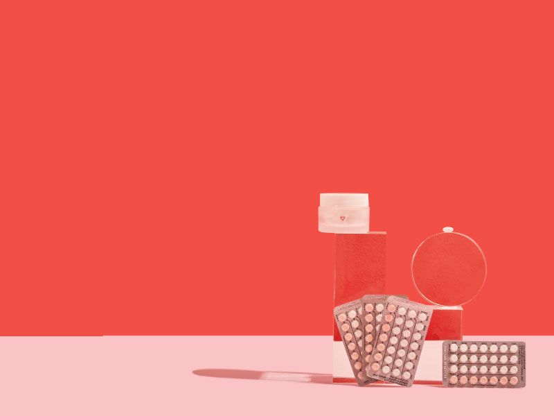 4 birth control packets and a small glass pill jar balanced on clear abstract shapes on a pink surface with a red background