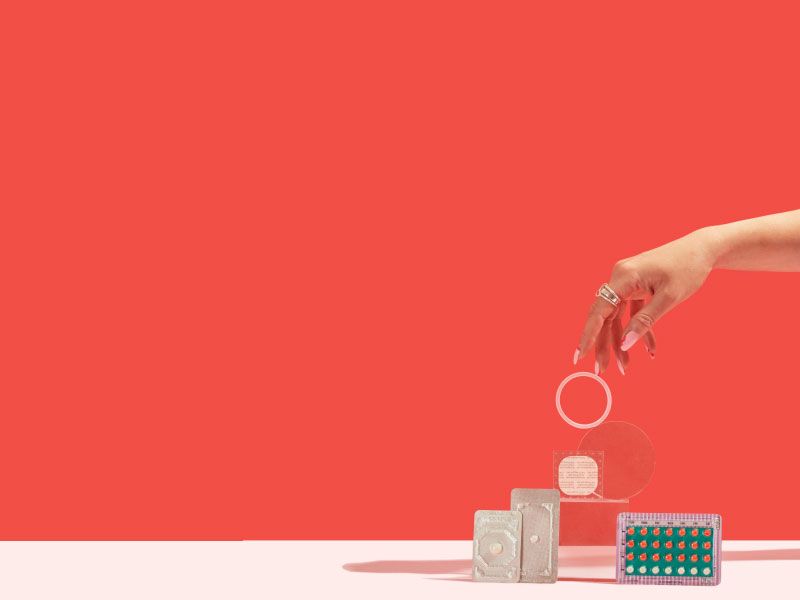 A woman's hand reaching for NuvaRing balanced on clear abstract shapes with the birth control patch, a birth control packet, Plan B, and Ella packets on a pink surface with a red background