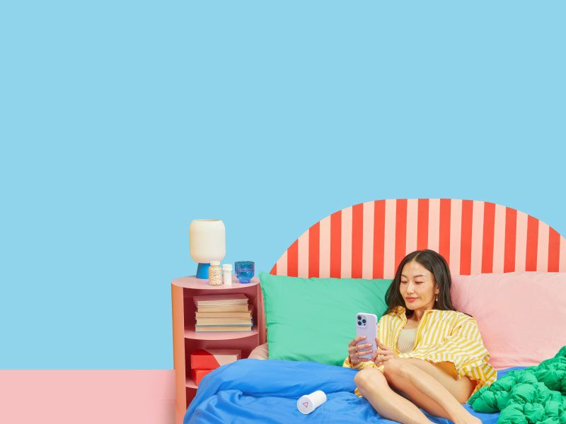 A woman laying on a bed with colorful bedding using a mobile phone next to a nightstand with Wisp medication bottles