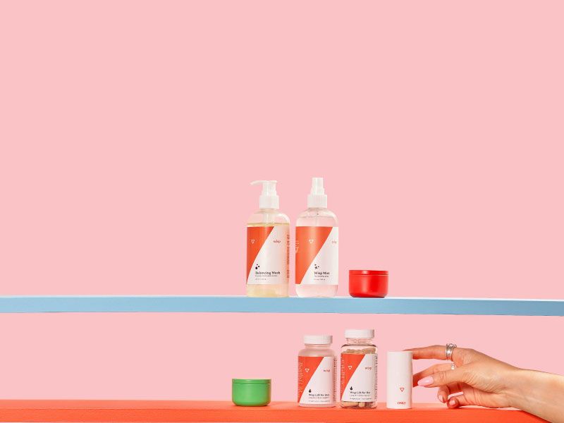 A variety of Wisp products on 3 colorful shelves with miscellaneous containers, a pink razor and a woman's hand reaching for Wisp OMG Cream in front of a pink background