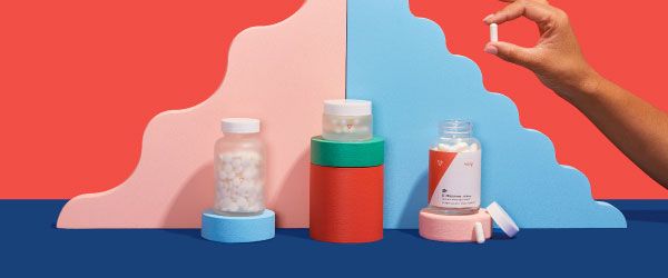 A person's hand is holding a pill next to various Wisp products balanced on colorful abstract shapes on a blue surface with a red background