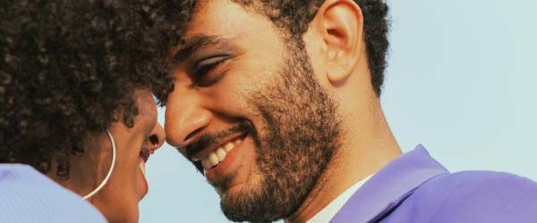 A woman and man with black curly hair and colorful clothing are looking into each other's eyes while smiling