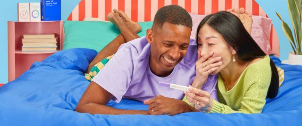 A man and woman wearing bright colored clothes are laying on a bed and smiling at each other while holding a pregnancy test