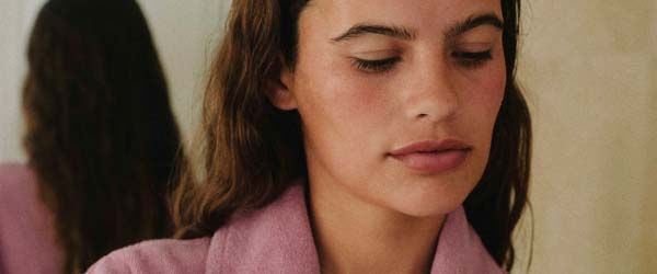 A woman with wavy hair wearing a pink robe is opening a sheet mask