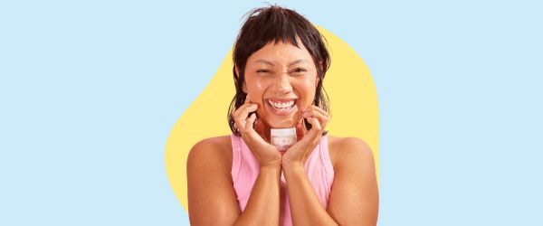 A smiling woman wearing a pink tank top is holding a Wisp glass jar under her chin in front of a light blue and yellow background