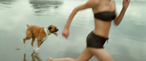A woman wearing a black swimsuit is running on the beach with her dog