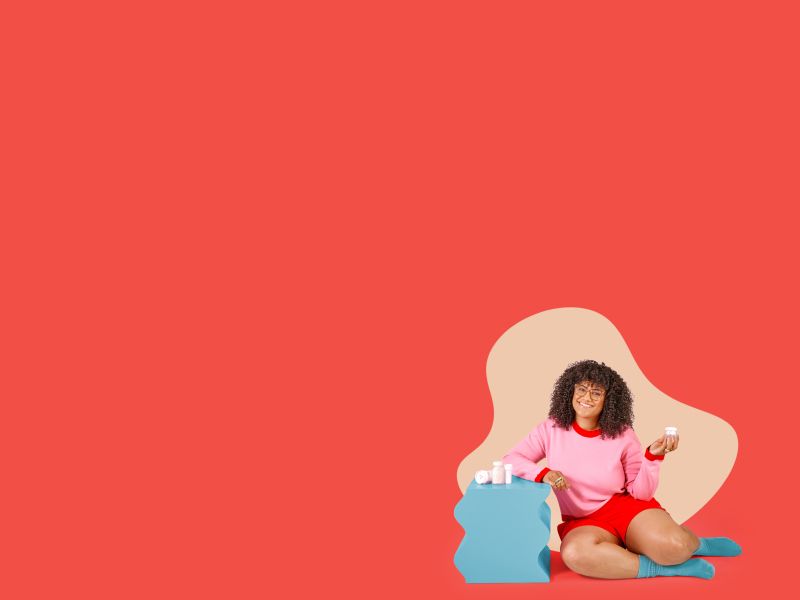 A woman wearing a pink sweater and red shorts is smilling and holding a Wisp glass pill jar while sitting next to a teal nightstand with various Wisp products on it