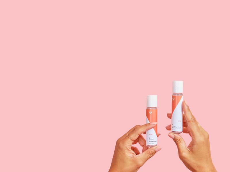 Two hands holding Wisp Original Harmonizing Lube and Wisp Toy-Safe Harmonizing Lube in front of a pink background