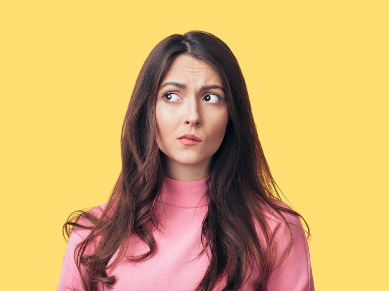 A woman with brown hair wearing a pink shirt has a confused expression in front of a yellow background