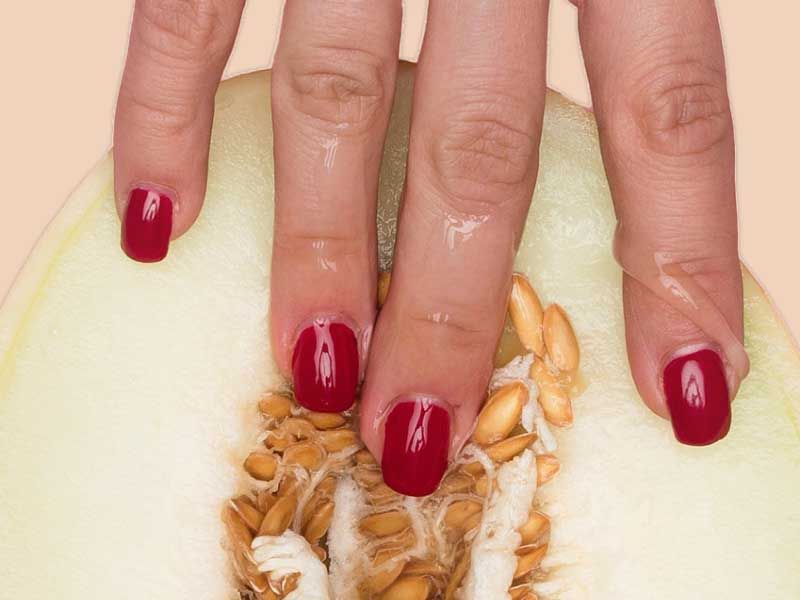 Hand with red nail polish touching the seeds of a sliced melon against a plain background.