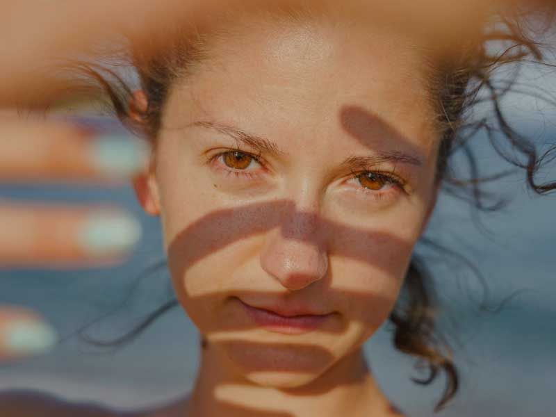 Close-up of a woman's eyes and forehead, with wet hair spread around her head, lit by warm sunlight casting soft shadows across her face.