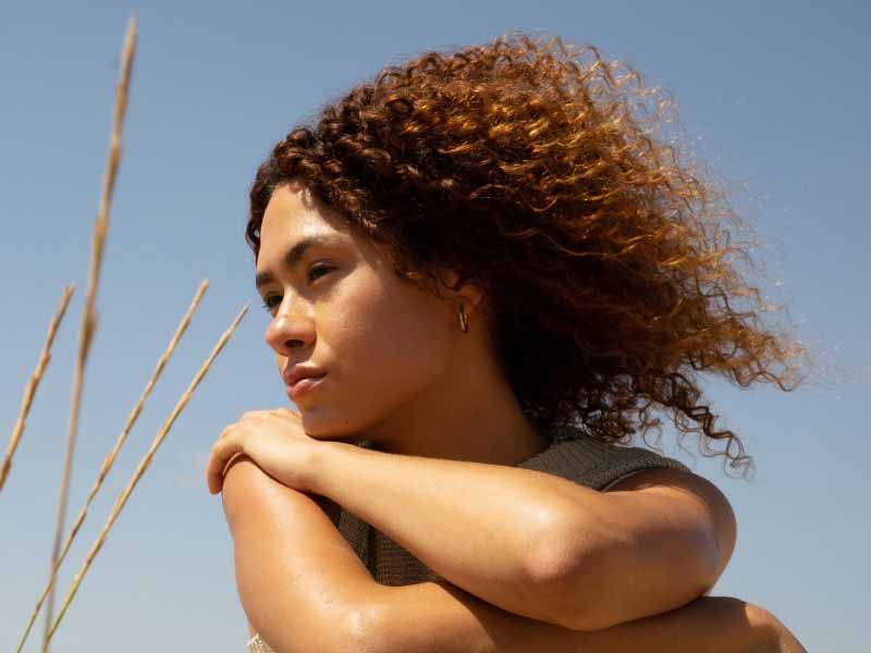Person with curly hair resting arms on knees, looking into the distance in a field