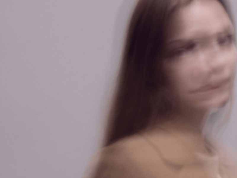 Blurred portrait of a young woman with long brown hair against a gray background.