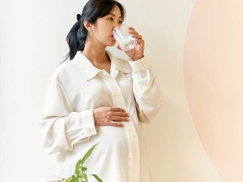 A pregnant woman wearing a white button up shirt with one hand on her belly while drinking a glass of water