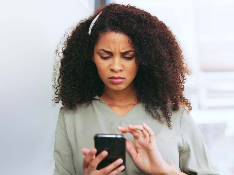 A woman wearing a gray shirt is looking at a mobile phone with a confused expression on her face