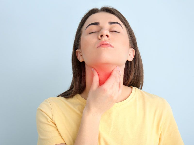 A woman wearing a yellow shirt has her hand on her throat in front of a light blue background