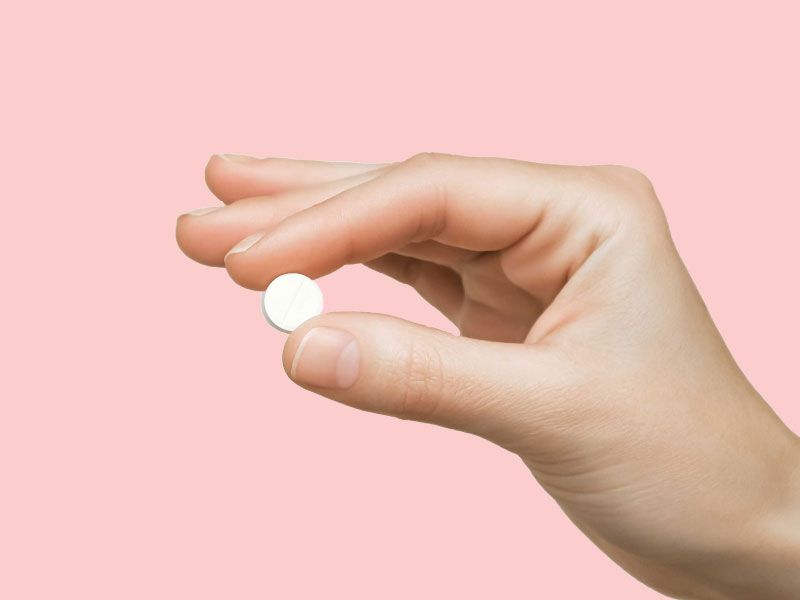 A person's hand holding a small round white pill in front of a pink background