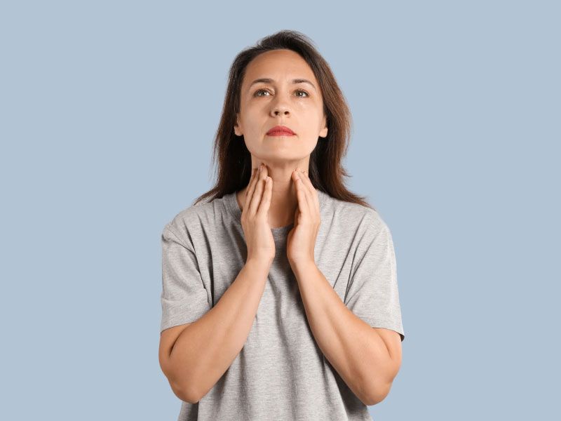 A woman wearing a gray shirt has her hands on her neck checking her thyroid