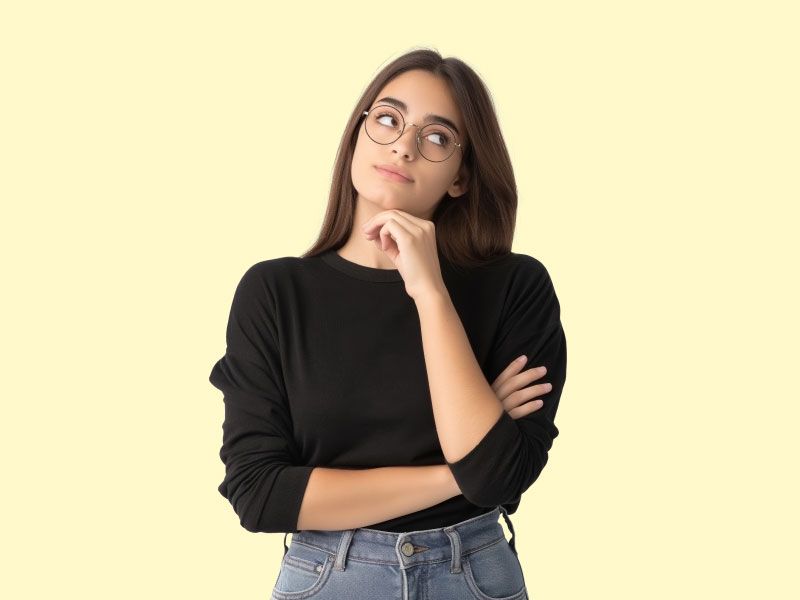 A woman wearing a black sweater and gray jeans is looking pensive in front of a yellow background