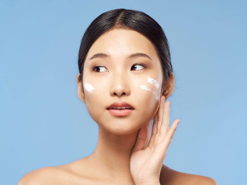 A pretty woman with glowing skin is applying tretinoin cream to her face in front of a blue background
