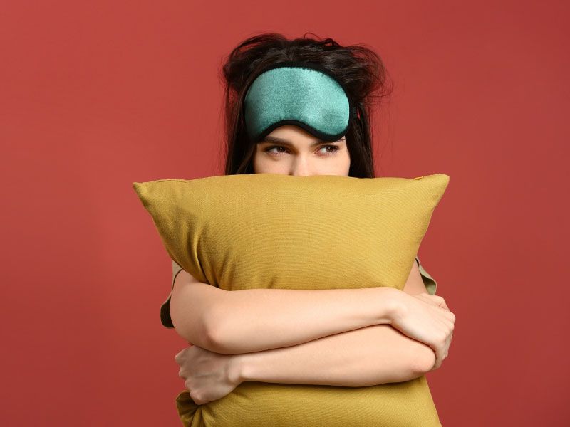 A woman wearing a green sleep mask and hugging a yellow pillow in front of a dark red background