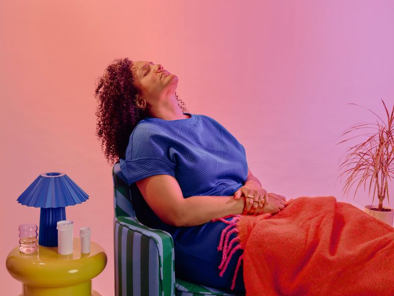 A woman wearing a blue shirt is sitting on a striped sofa with her head back in discomfort next to a yellow nightstand with a blue lamp and various Wisp medication vials
