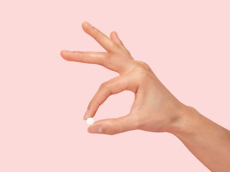 A hand holding a small round white pill between the index finger and thumb with a pink background