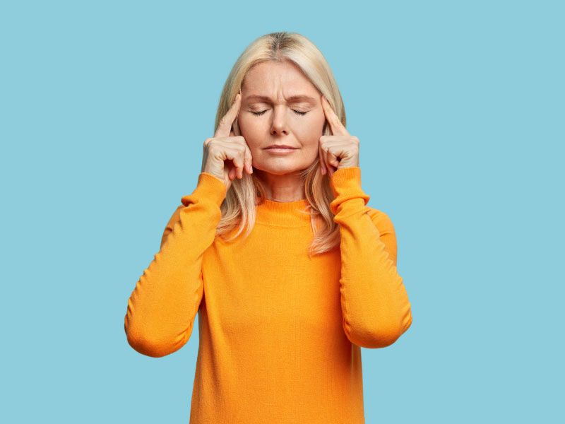 A woman with blonde hair is wearing a yellow sweater with her fingers on her temples in discomfort in front of a blue background