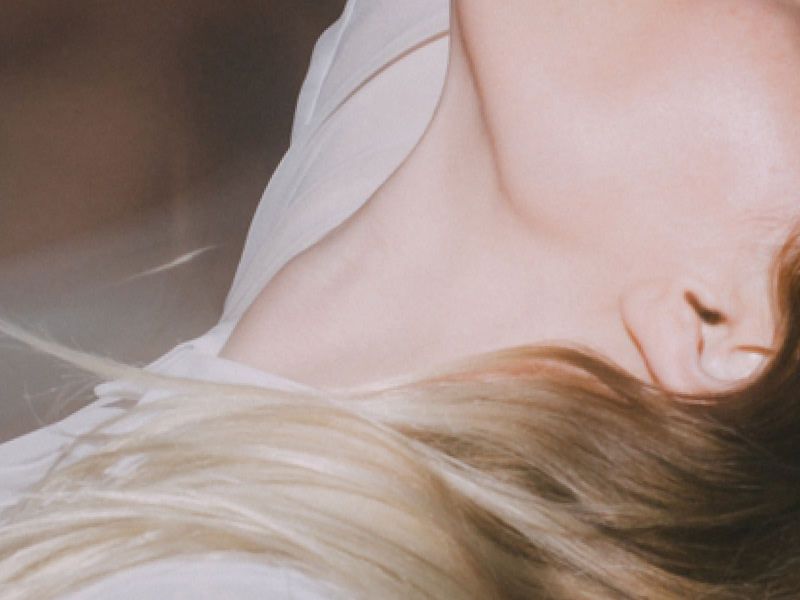 image of a woman's left side profile with a cast of light on her cheek, neck and blonde hair wearing a white shirt and a blurry image of a mans neck and chest in the background