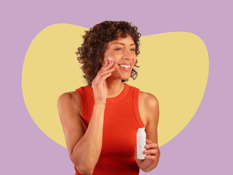 A smiling woman wearing a red tank top is applying Topical Spironolactone cream to her face