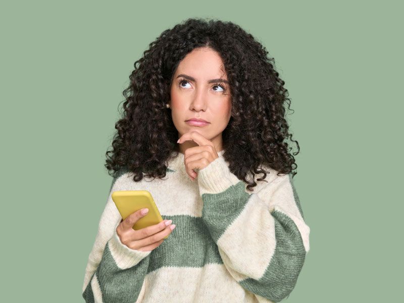 A woman wearing a striped green and beige sweater has her hand on her chin in contemplation while looking at a mobile phone